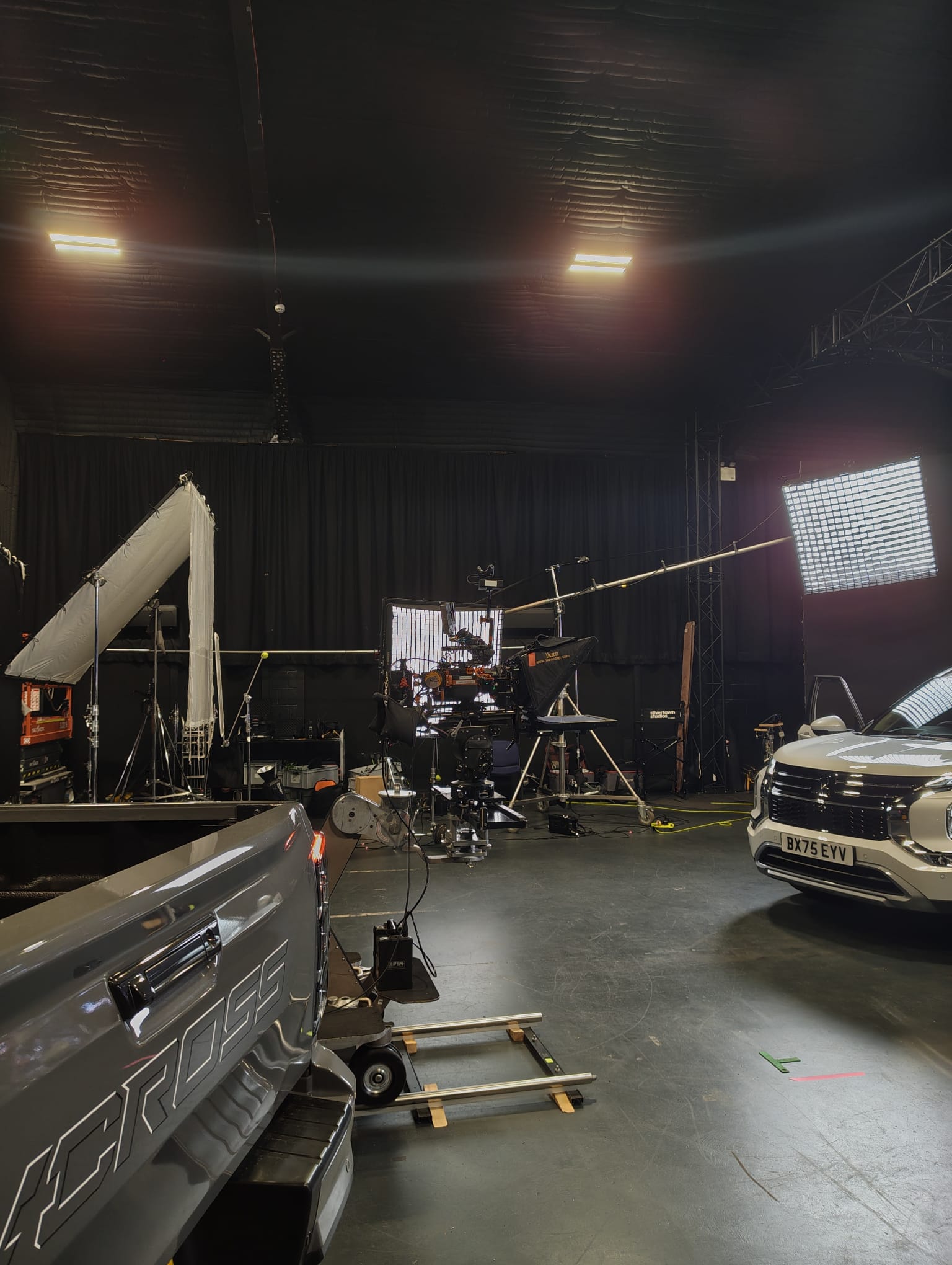A studio set for a vehicle commercial — two cars lit with diffusion frames, a camera dolly on track and lighting rigs.