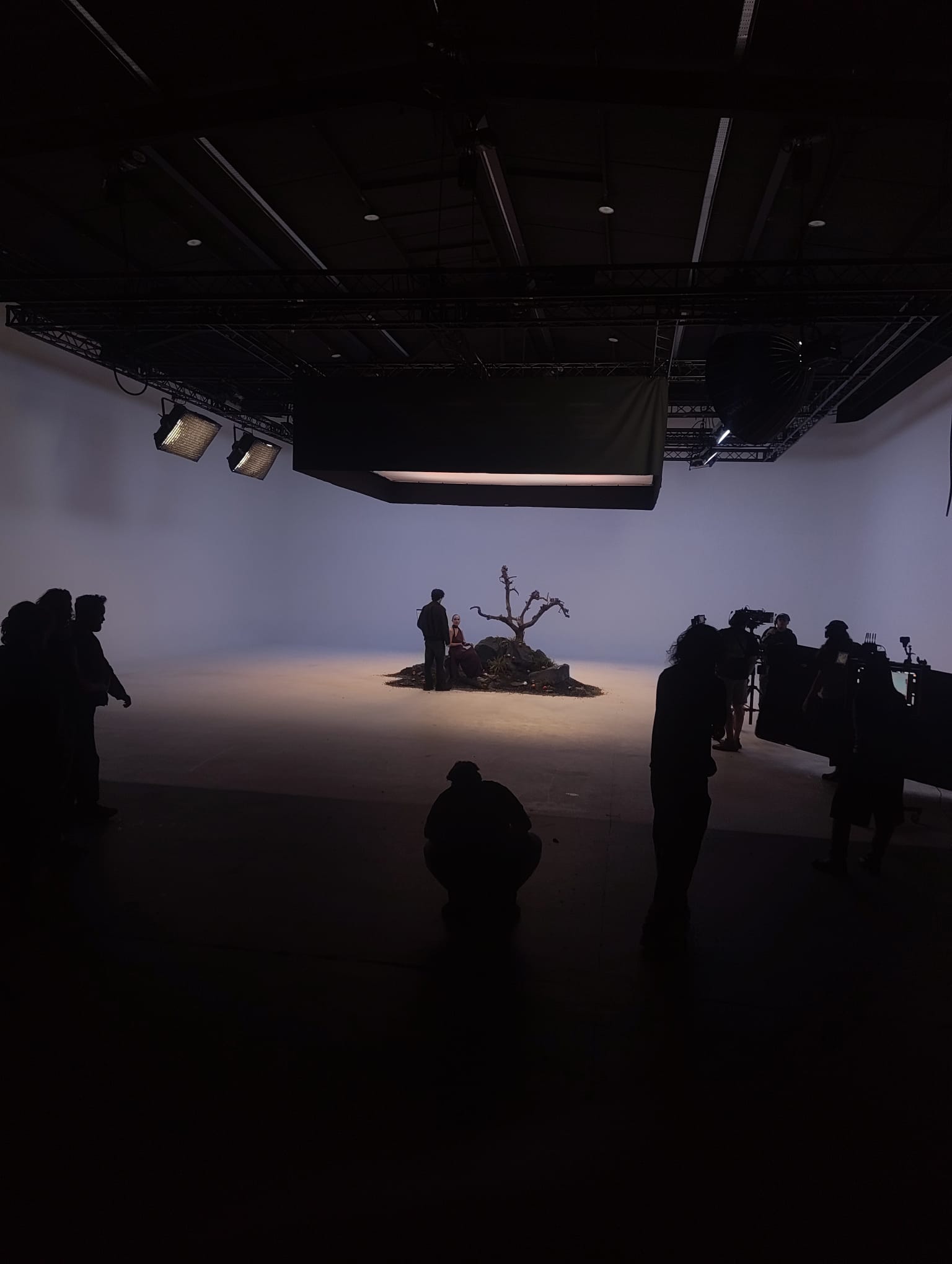 A darkened cyclorama stage with silhouetted crew around a single lit set piece — a bare tree on a small island of earth.